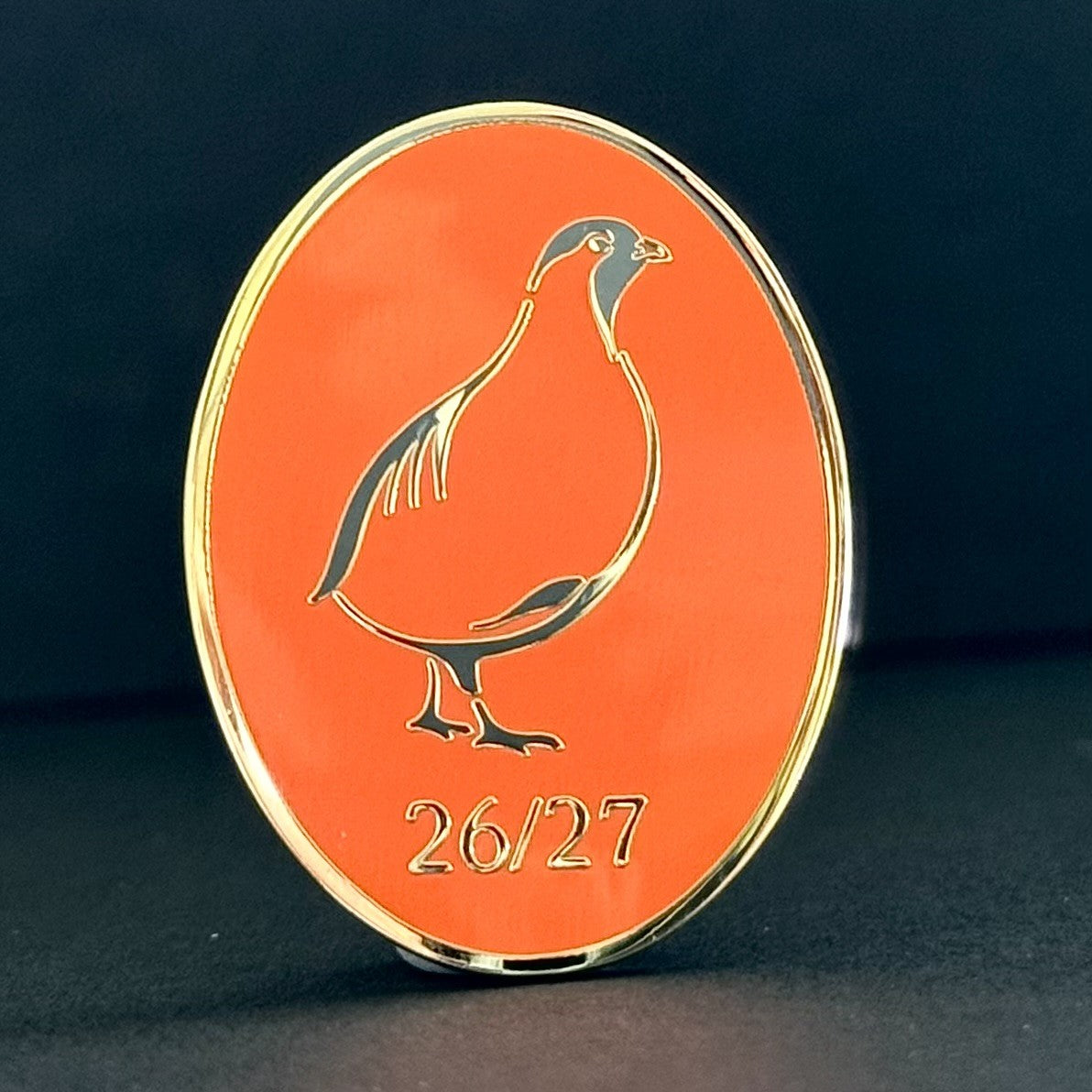An oval orange collectible coin or medallion with a gold-plated border, featuring a gold bird silhouette and the text '26/27' at the bottom, set against a dark background.